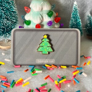 May include: A gold-tone enamel pin shaped like a green Christmas tree with white and yellow sprinkles. The pin is on a gray background with the text "tabauble.com".
