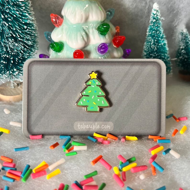 May include: A gold-tone enamel pin shaped like a green Christmas tree with white and yellow sprinkles. The pin is on a gray background with the text "tabauble.com".