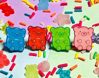 May include: Four gummy bear shaped pins in blue, red, green, and pink. The pins are arranged in a row on a colorful background with sprinkles and other candy.