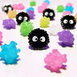 May include: A selection of colourful, candy-like objects, including black soot sprites with white eyes. The other objects are various colours, including pink, purple, blue, green, and yellow. The objects are scattered on a white surface.