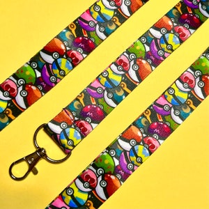 May include: A colourful lanyard with a repeating pattern of Poke Balls in various colours and designs. The lanyard has a silver metal clip.