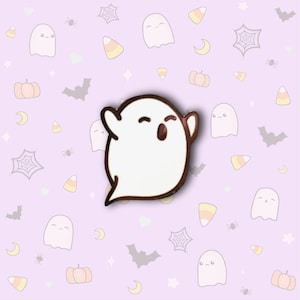 May include: White enamel pin of a cartoon ghost with a brown outline. The ghost has a wide smile and its eyes are closed. The pin is on a purple background with a pattern of bats, pumpkins, and candy corn.
