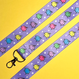 May include: Purple lanyard with a repeating pattern of colorful cartoon characters and white stars. The lanyard has a silver-colored metal clasp. The background is a solid yellow.