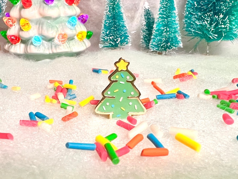 May include: A green and gold Christmas tree pin with white and yellow sprinkles. The pin is on a white surface with colorful sprinkles scattered around it.
