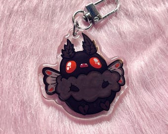 May include: A clear acrylic keychain featuring a cartoon moth design. The moth is primarily black with red eyes and accents, and has a fluffy body and wings. The keychain has a silver clasp for attachment.