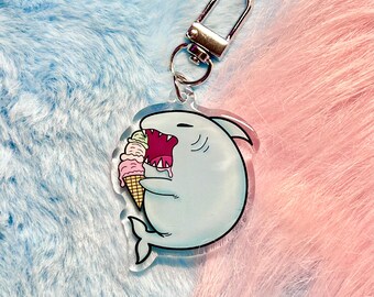 May include: A blue shark keychain with a silver key ring. The shark is holding a multi-colored ice cream cone in its mouth.