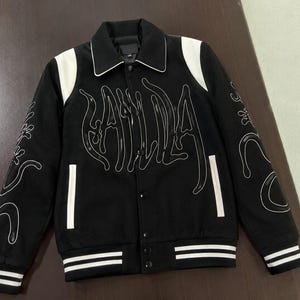 以下が含まれることがあります： 黒のバーシティジャケット。白いショルダーパネル、折り返し襟、スナップボタン留めが特徴。フロントと袖には、光沢のある黒の立体的な文字と、白い輪郭線で描かれた抽象的なグラフィックデザインが施されています。