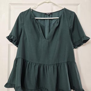 May include: A dark green short-sleeved blouse featuring a deep V-neckline, a tiered peplum-style hem, and ruffled edges on the sleeves and bottom. The garment includes a Madewell brand label at the inner neckline and is displayed on a white hanger.
