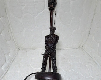 May include: A dark brown metal table lamp base featuring a sculpted figure of a golfer in a cap holding a golf club. The lamp includes a vertical rod, a pull chain switch, and a standard light bulb socket at the top with a black power cord.