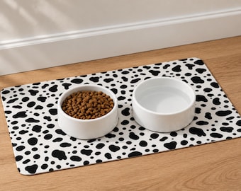 May include: A rectangular pet feeding mat with a black and white cow print pattern. Two round white ceramic bowls sit on the mat, one filled with brown dry kibble and the other filled with clear water, creating a functional pet dining set.