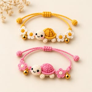 Puede incluir: Dos pulseras ajustables de ganchillo con pequeños dijes de tortuga con caras sonrientes. Una pulsera es amarilla con flores blancas y cascabeles dorados, y la otra es rosa con flores a juego y cascabeles dorados, ambas con extremos anudados.