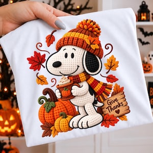 Peut inclure: Un t-shirt blanc avec un motif de chien de style crochet portant un bonnet et une écharpe en tricot, tenant une tasse avec de la crème fouettée. Le design inclut des citrouilles orange, des feuilles d'automne et un panneau en bois marqué Give Thanks.