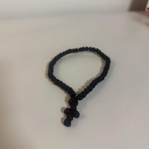 May include: A black knotted cord bracelet featuring a small, integrated cross-shaped charm made from the same black cord material. The bracelet is constructed from a series of uniform, tightly tied knots forming a continuous circular band.