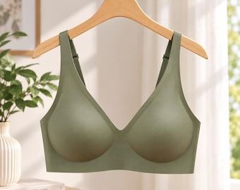 May include: A sage green wireless bra with moulded cups, a deep V-neckline, and wide shoulder straps. The garment features a seamless construction and is displayed on a light-coloured wooden hanger against a bright, neutral background.
