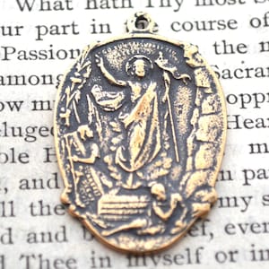 May include: Antique gold-colored religious pendant with an oval shape. The pendant features a detailed relief of a figure with outstretched arms, surrounded by other figures and text. The pendant has a small loop at the top for hanging.
