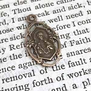 May include: Silver pendant with a detailed design featuring a religious figure and floral accents. The pendant is oval-shaped and has a loop at the top for attaching to a chain.