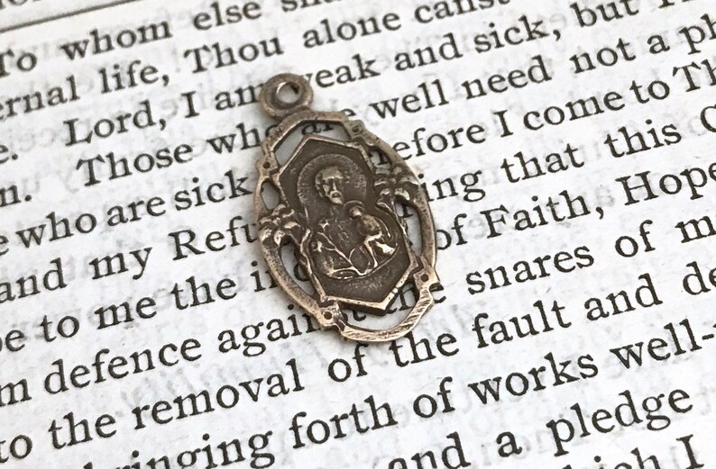 May include: Silver pendant with a detailed design featuring a religious figure and floral accents. The pendant is oval-shaped and has a loop at the top for attaching to a chain.