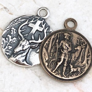 May include: Two silver tone pendants with a religious theme. One pendant features a deer with antlers and a cross. The other pendant features a man with a spear and a deer.
