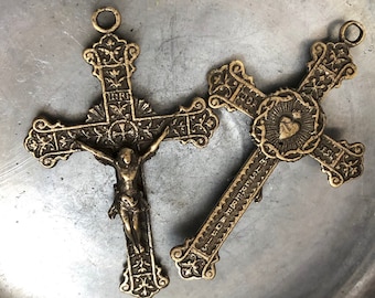 May include: Two antique brass crucifixes. One depicts Jesus on the cross with the letters INRI above. The other cross has a heart in the center and text along the arms. Both crosses have ornate detailing and a loop at the top for hanging.