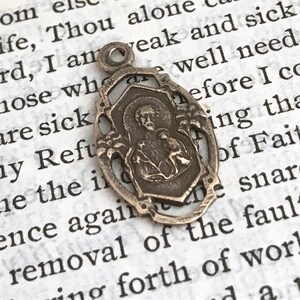 May include: A silver-toned metal pendant featuring a religious image of a person with a halo. The pendant is attached to a loop at the top and has a detailed design with intricate floral accents.