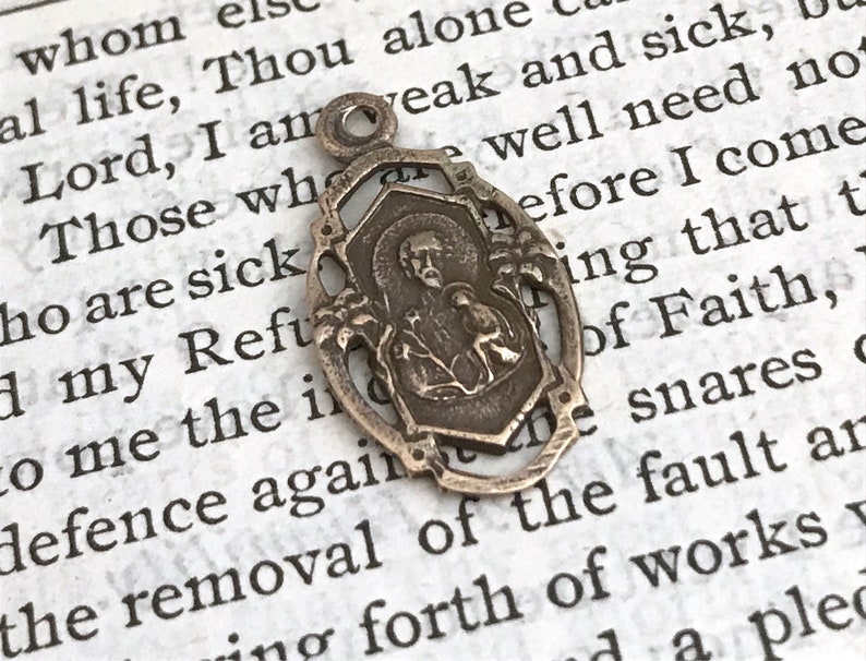 May include: A silver-toned metal pendant featuring a religious image of a person with a halo. The pendant is attached to a loop at the top and has a detailed design with intricate floral accents.