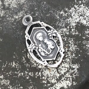 May include: A silver charm with a detailed design featuring a religious figure. The charm has a loop at the top for attaching to a necklace or bracelet.