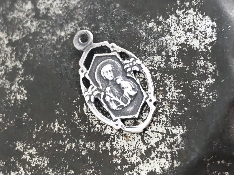 May include: A silver charm with a detailed design featuring a religious figure. The charm has a loop at the top for attaching to a necklace or bracelet.