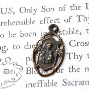 May include: A small, antique, bronze-colored pendant featuring a religious image of a person holding a child. The pendant is on a white background with text.