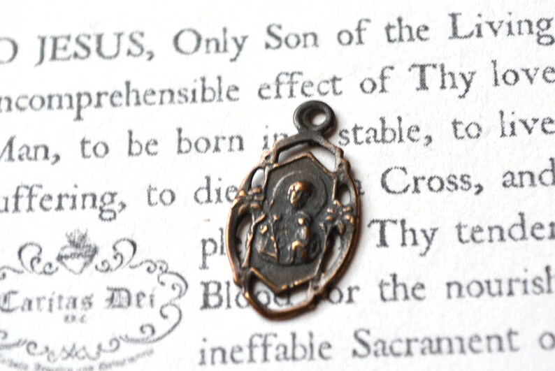 May include: A small, antique, bronze-colored pendant featuring a religious image of a person holding a child. The pendant is on a white background with text.