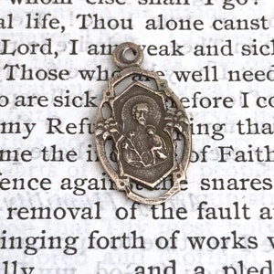 May include: Silver pendant featuring a religious image of a saint with a bird. The pendant is oval shaped with a decorative border. The pendant is on a white background with text.