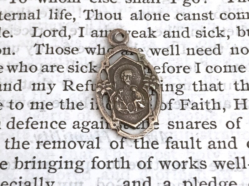 May include: Silver pendant featuring a religious image of a saint with a bird. The pendant is oval shaped with a decorative border. The pendant is on a white background with text.