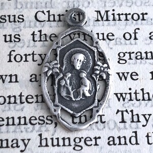 May include: Silver pendant featuring a religious image of a saint holding a bird. The pendant is oval shaped with a detailed design and a loop at the top for hanging.