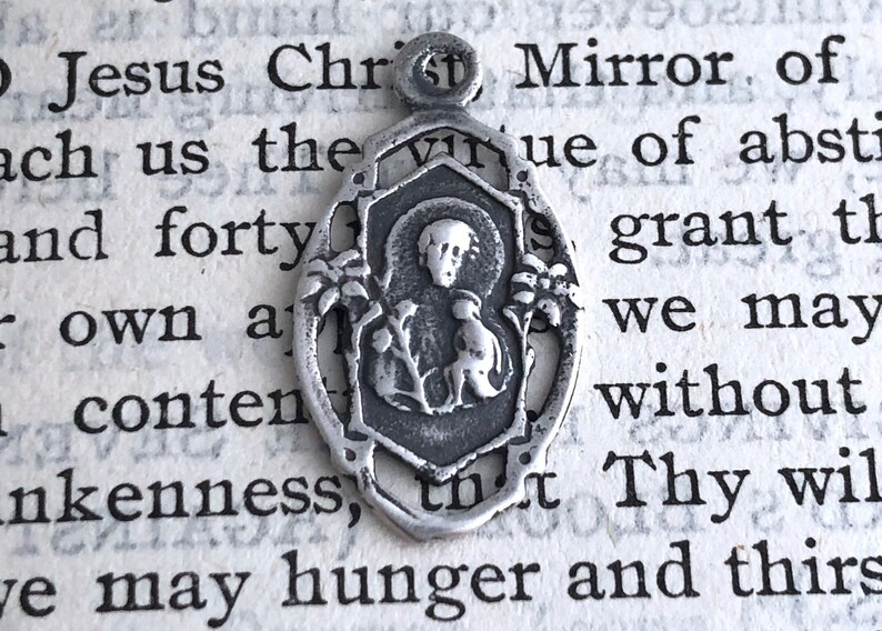 May include: Silver pendant featuring a religious image of a saint holding a bird. The pendant is oval shaped with a detailed design and a loop at the top for hanging.