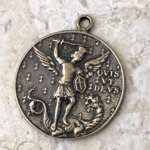May include: A bronze pendant featuring a depiction of Saint Michael the Archangel slaying a dragon. The pendant is circular and has a loop at the top for hanging. The inscription on the pendant reads "QUIS UT DEUS".