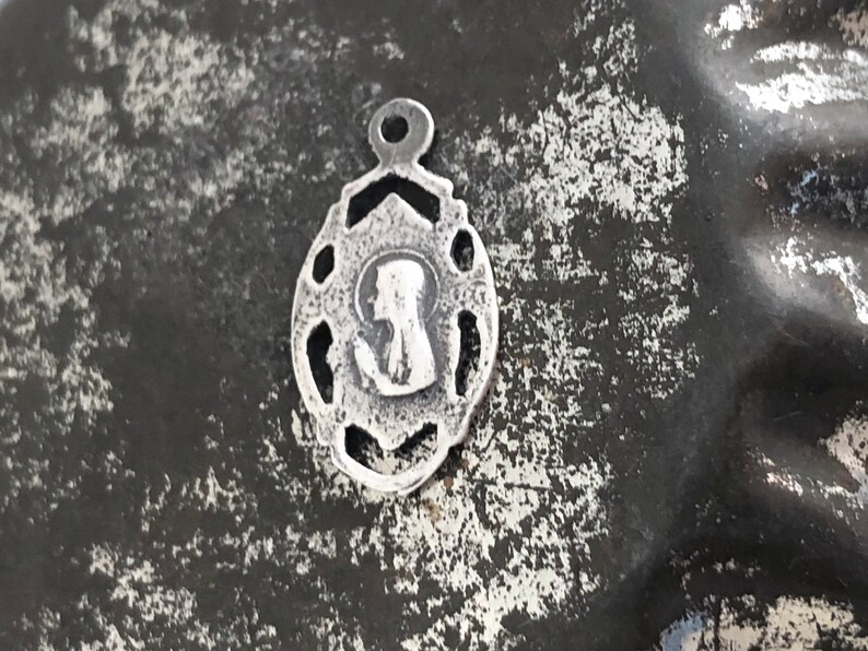 May include: A silver charm with a detailed design featuring a silhouette of a woman's head. The charm has a loop at the top for attaching to a necklace or bracelet.