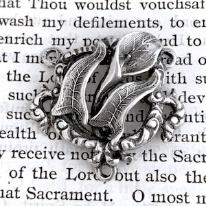 May include: A silver charm with a floral design. The charm is in the shape of two leaves with a flower in the center. The charm is detailed and intricate.