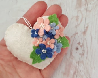 May include: A white felt heart-shaped ornament with a cluster of pink and blue felt flowers and green felt leaves.