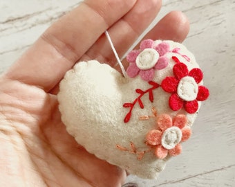 May include: A white felt heart with embroidered flowers in pink, red, and orange. The flowers have white centers and are surrounded by green stems.