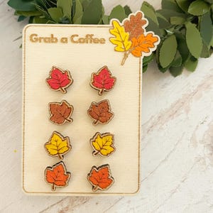 May include: Wooden leaf-shaped earrings in red, brown, yellow, and orange, displayed on a wooden card with the text "Grab a Coffee." The earrings are arranged in pairs, showcasing fall colors and a seasonal theme.