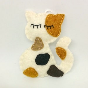 May include: A white felt cat with brown, orange, and black felt patches. The cat has closed eyes and a stitched smile.