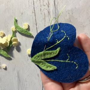 Puede incluir: Un coraz&oacute;n de fieltro azul con hojas y tallos bordados en verde, y una aguja e hilo. Flores y hojas de fieltro amarillas y verdes dispersas, sugiriendo un proyecto de manualidades.