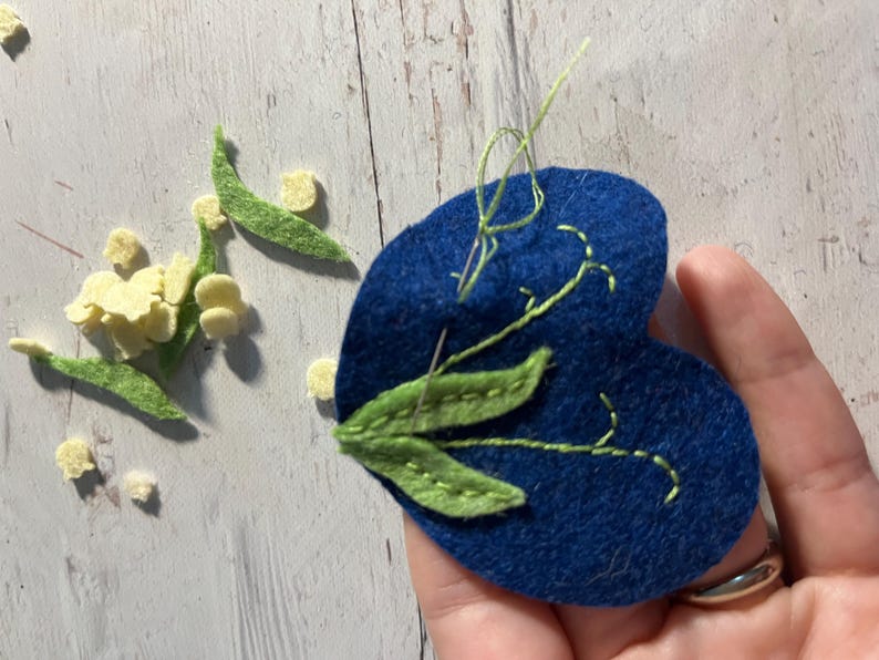 Puede incluir: Un coraz&oacute;n de fieltro azul con hojas y tallos bordados en verde, y una aguja e hilo. Flores y hojas de fieltro amarillas y verdes dispersas, sugiriendo un proyecto de manualidades.