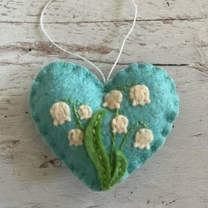 Puede incluir: Adorno en forma de coraz&oacute;n de fieltro turquesa con una cuerda blanca para colgar. Presenta flores de lirio de los valles bordadas con flores blancas en forma de campana y hojas verdes. Los bordes est&aacute;n cosidos con hilo turquesa.