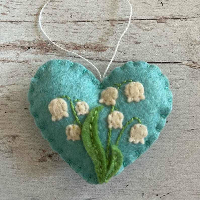 Puede incluir: Adorno en forma de coraz&oacute;n de fieltro turquesa con una cuerda blanca para colgar. Presenta flores de lirio de los valles bordadas con flores blancas en forma de campana y hojas verdes. Los bordes est&aacute;n cosidos con hilo turquesa.