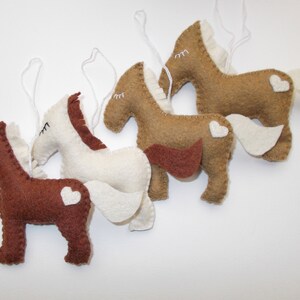 May include: Four felt horse ornaments with white hearts on their backs. The horses are brown, tan, white, and brown. They are all hanging from white strings.