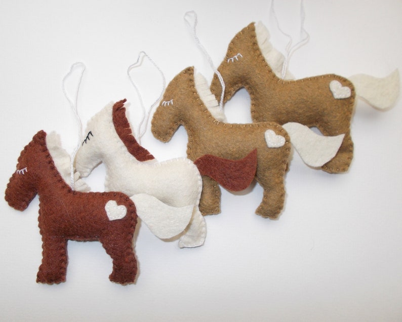 May include: Four felt horse ornaments with white hearts on their backs. The horses are brown, tan, white, and brown. They are all hanging from white strings.