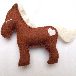 May include: A brown felt horse ornament with a white felt mane and tail. The horse has a small white heart on its back.