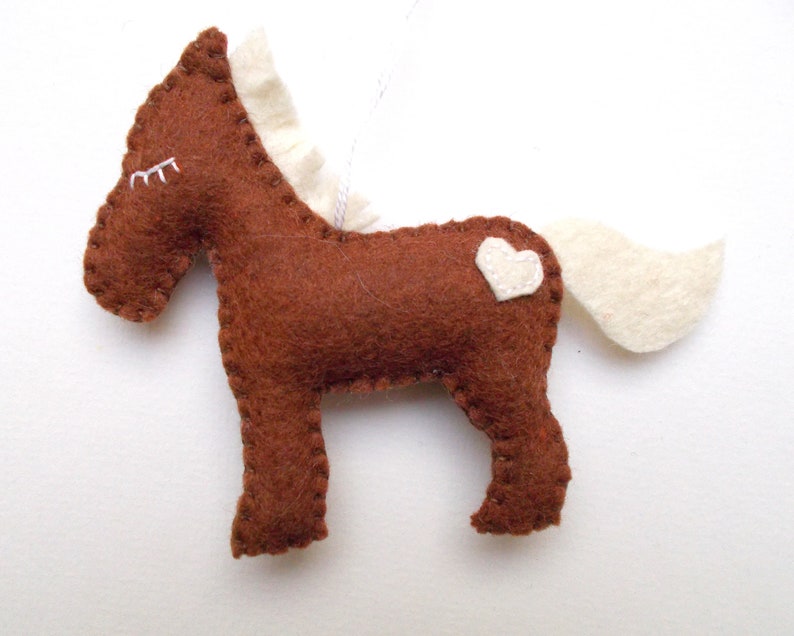 May include: A brown felt horse ornament with a white felt mane and tail. The horse has a small white heart on its back.