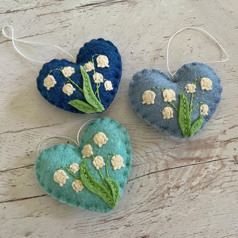 Puede incluir: Tres adornos en forma de coraz&oacute;n en tonos azules y turquesas, cada uno adornado con un dise&ntilde;o de lirios de los valles. Los adornos de fieltro presentan flores blancas y hojas verdes, con una cuerda blanca para colgar. Las costuras son visibles.