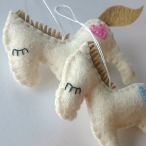 May include: Two white felt donkey ornaments with black eyes and brown felt manes. One donkey has a pink heart and the other has a blue heart.